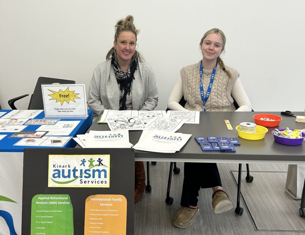 Two individuals sitting and smiling at an information booth about Kinark Autism Services