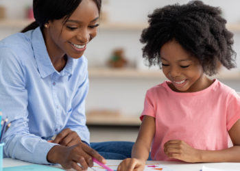 An adult female sits at a table with a female child looking at paperwork. Both people have dark skin and hair.